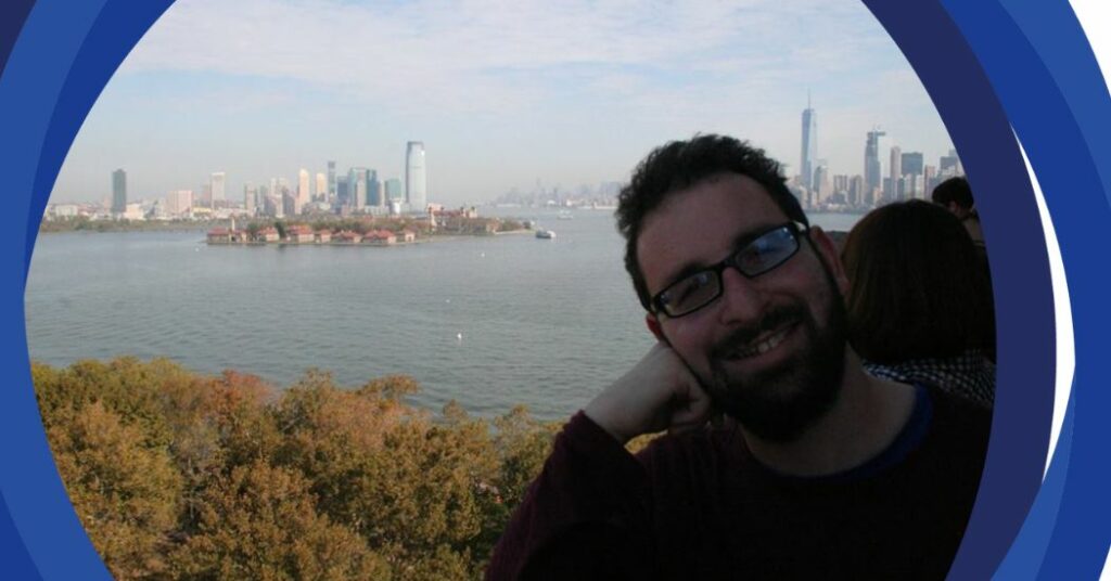 Simon smiling with a park, riverfront, and a city skyline with many skyscrapers in the background.