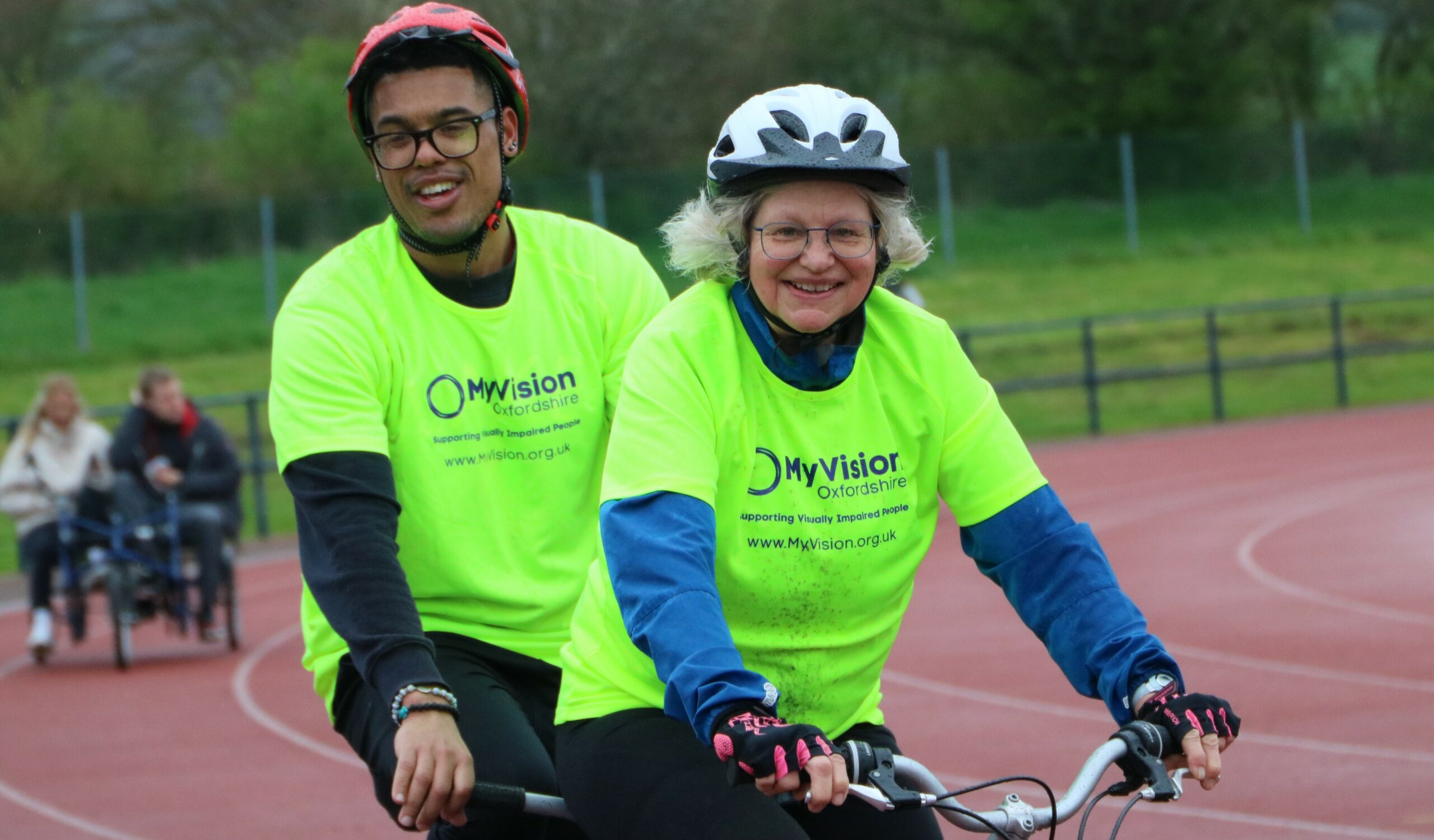 A photo of two people wearing MyVision t-shirts, riding a tandem bike and smiling at the camera