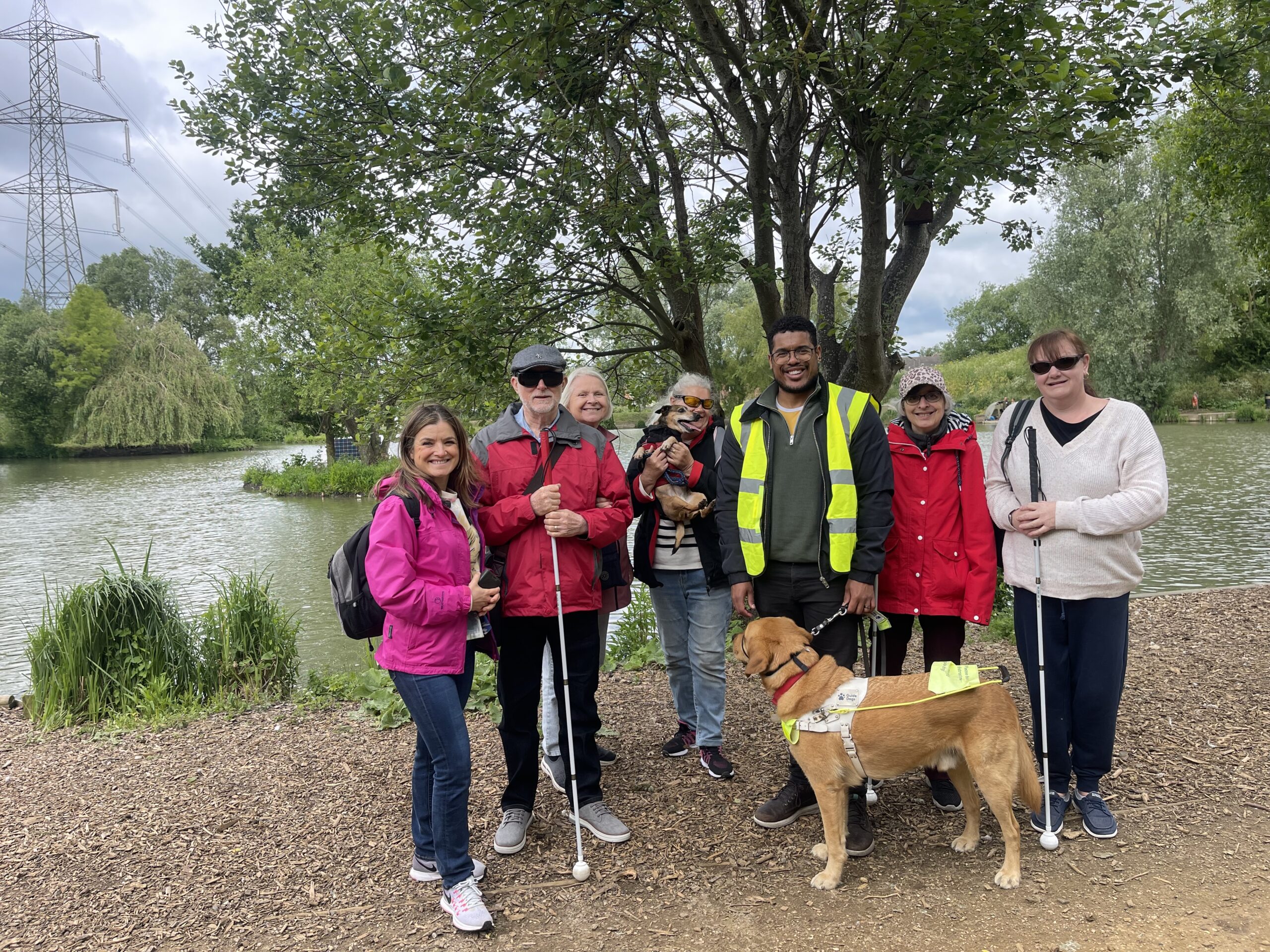 A group of people standing on a path by a lake. They are smiling at the camera. There is a guide dog too. 