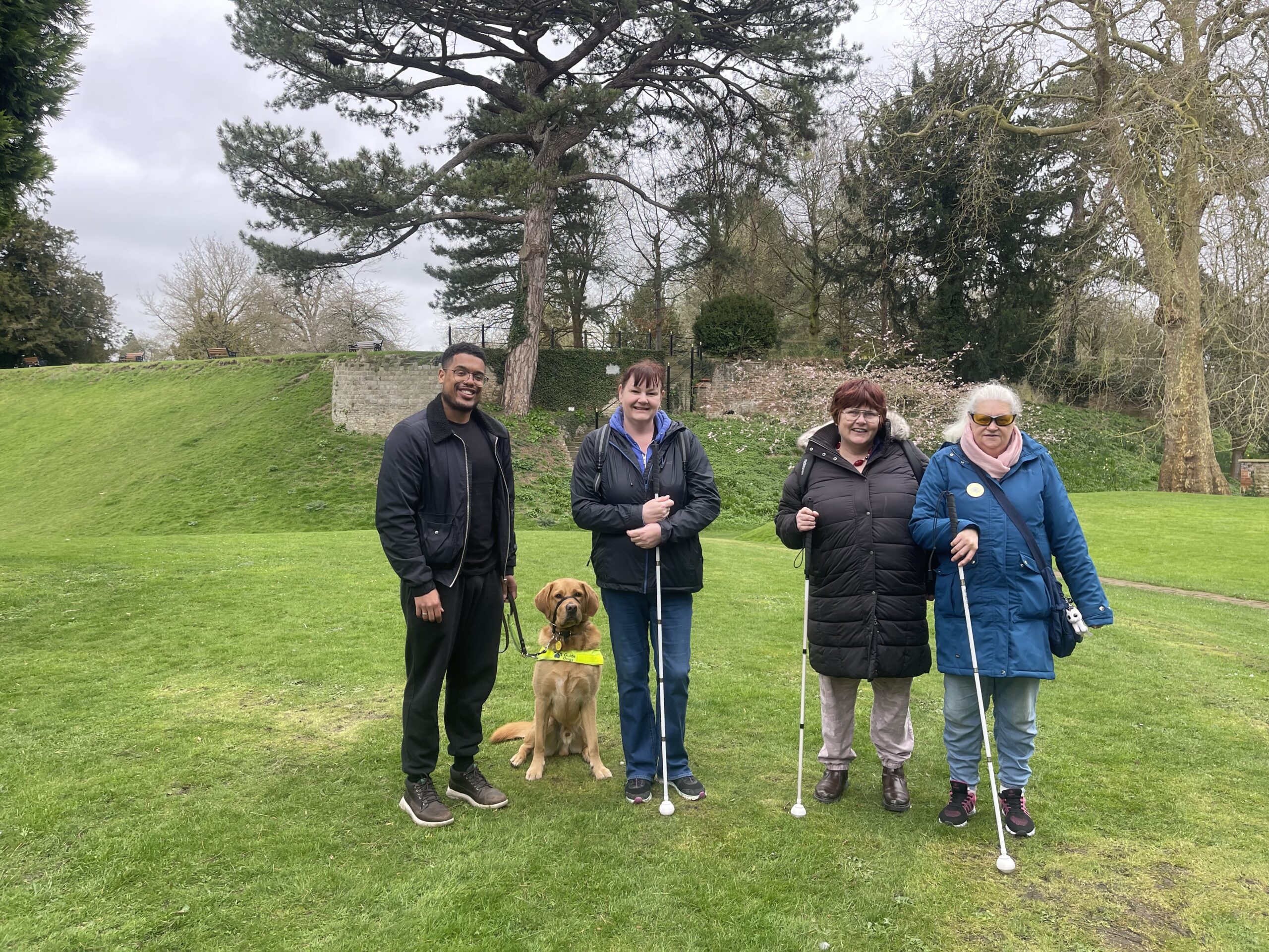 Four people standing on some grass smiling at the camera. There is a guide dog too