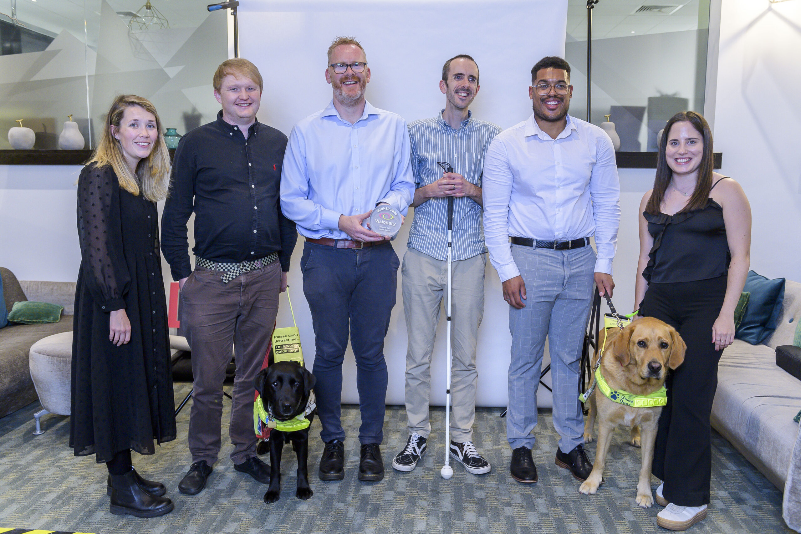 Six of the MyVision teams standing in a line smiling at the camera. The man in the middle is holding up the Innovate and Inspire award. There are also two guide dogs.