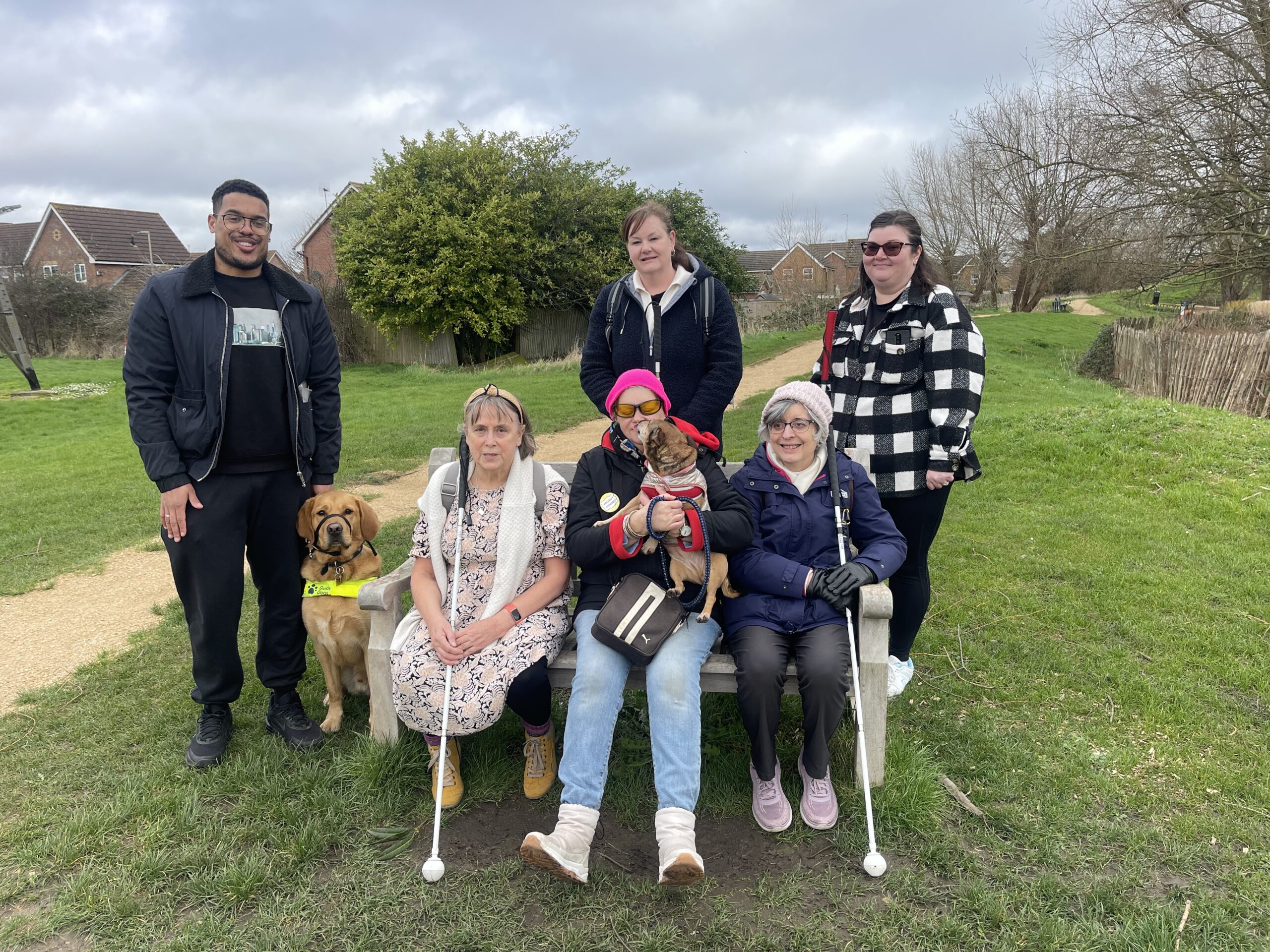 a group photo. There are three women sitting on a bench, one of them has a dog. there are three people stood behind them, one of them has a guide dog.