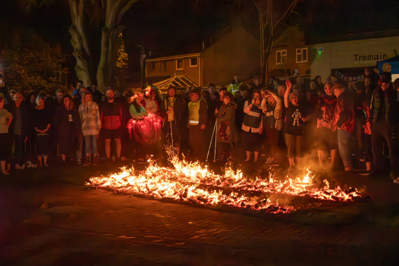 A photo of a firewalk. There is a long path of coals, with flames either side, and a group of people standing around. Some of the people have canes.